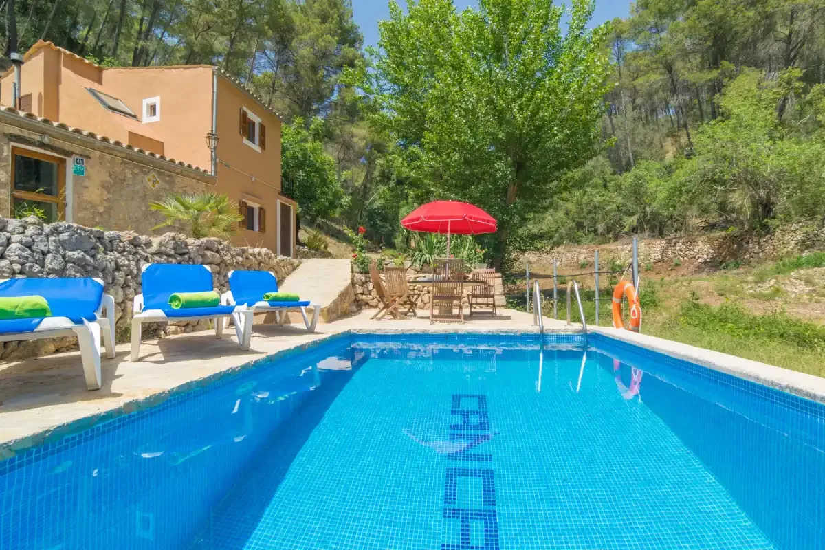 Una piscina al aire libre junto a una casa, con asientos y una sombrilla roja.