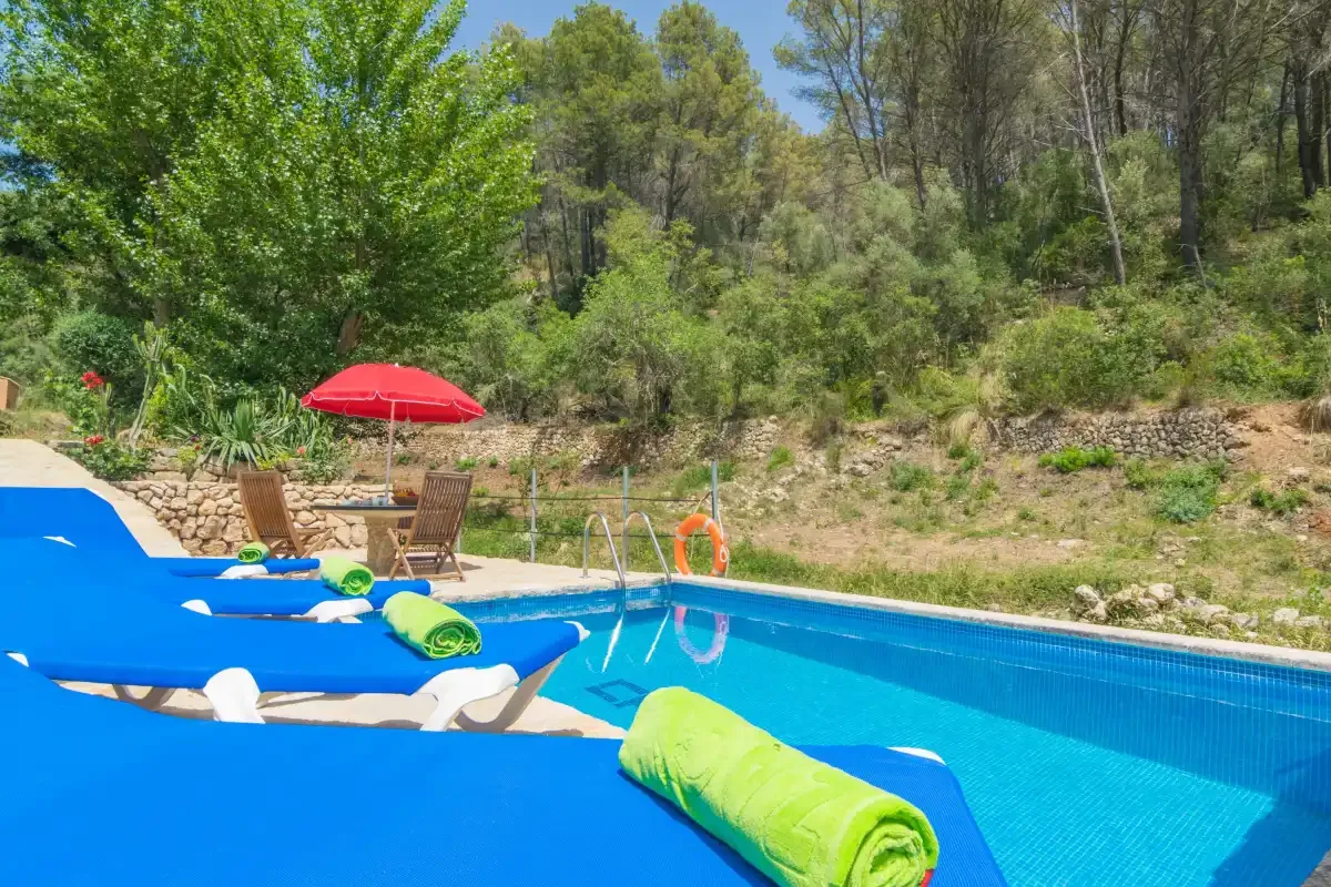 Una piscina al aire libre con tumbonas azules junto a una ladera arbolada.