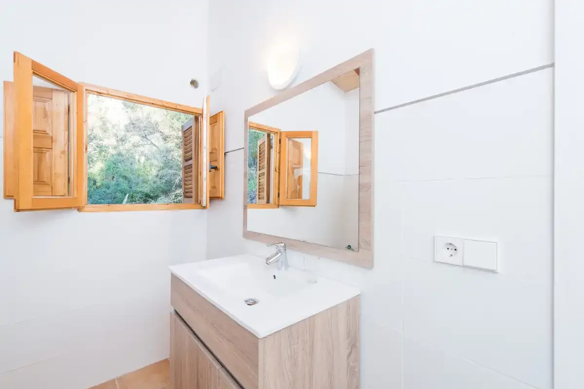 Un baño luminoso con lavabo, espejo y ventana de madera abierta.