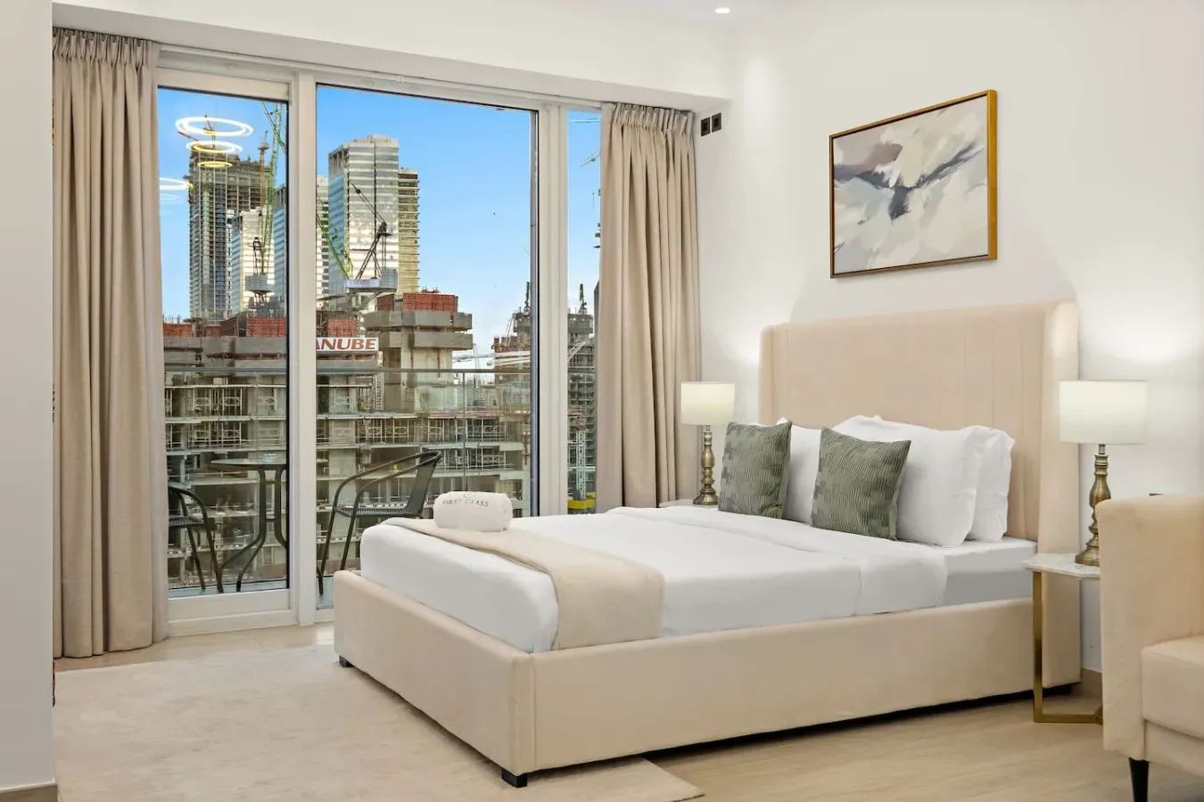 Un dormitorio luminoso con una cama tendida junto a grandes ventanas con vistas a edificios de la ciudad.