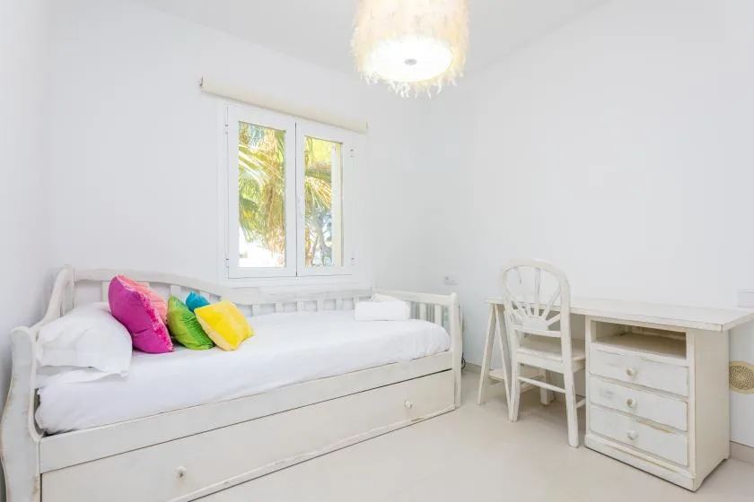 Un dormitorio luminoso con una cama blanca, cojines de colores y un escritorio junto a la ventana.