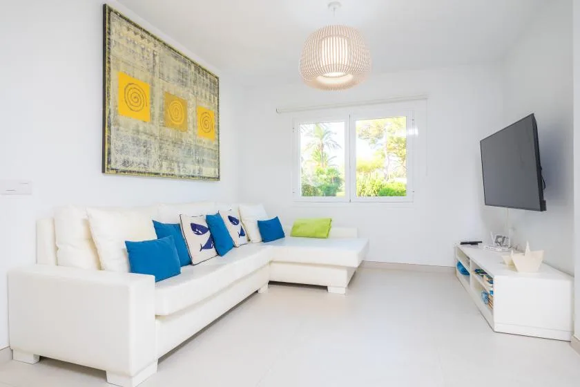 Una sala luminosa con asientos blancos, cojines de colores y un televisor en la pared.