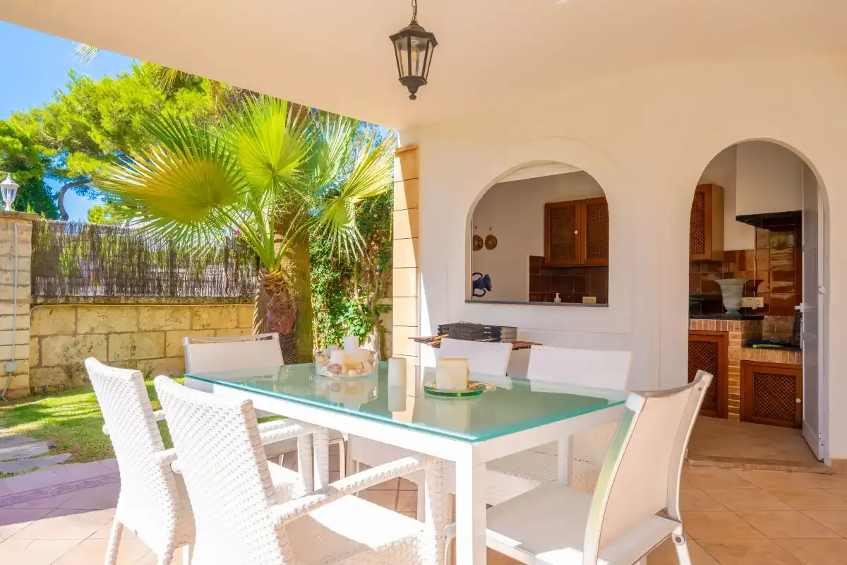 Un comedor cubierto en el patio, con asientos blancos y una mesa de vidrio, da a una palmera.