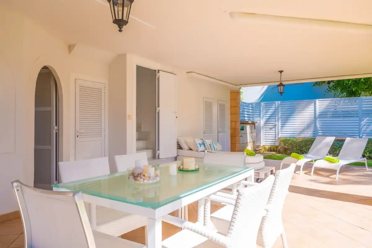 Un patio cubierto con una mesa de comedor de vidrio y asientos blancos.