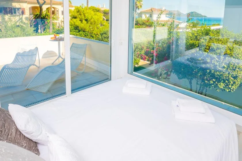 Un dormitorio con una cama blanca junto a ventanales con vistas a la vegetación y al mar.