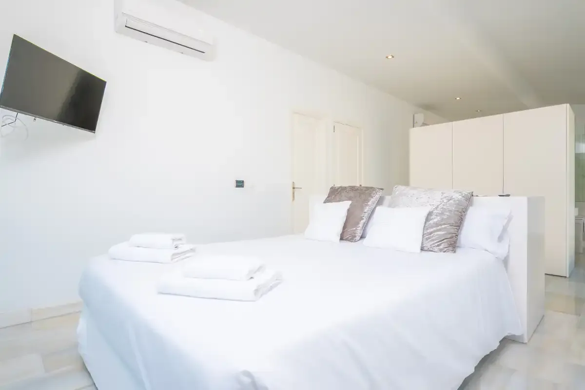 Un dormitorio luminoso con una cama tendida y un televisor en la pared.
