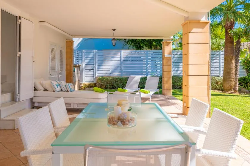 Un patio cubierto tiene una mesa de comedor con tapa de vidrio y asientos junto al jardín.