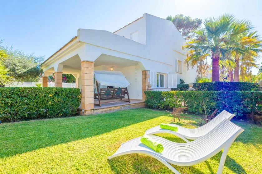 Una casa blanca con patio cubierto, césped y asientos reclinables al aire libre.