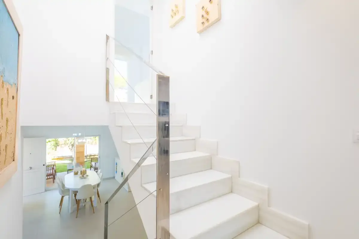 Un interior luminoso muestra una escalera blanca junto a una barandilla de cristal y un comedor abajo.