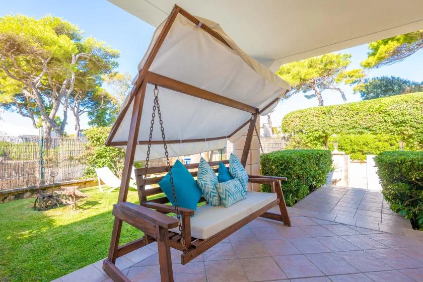 Un patio cubierto tiene un columpio de madera con cojines azules junto a un jardín.