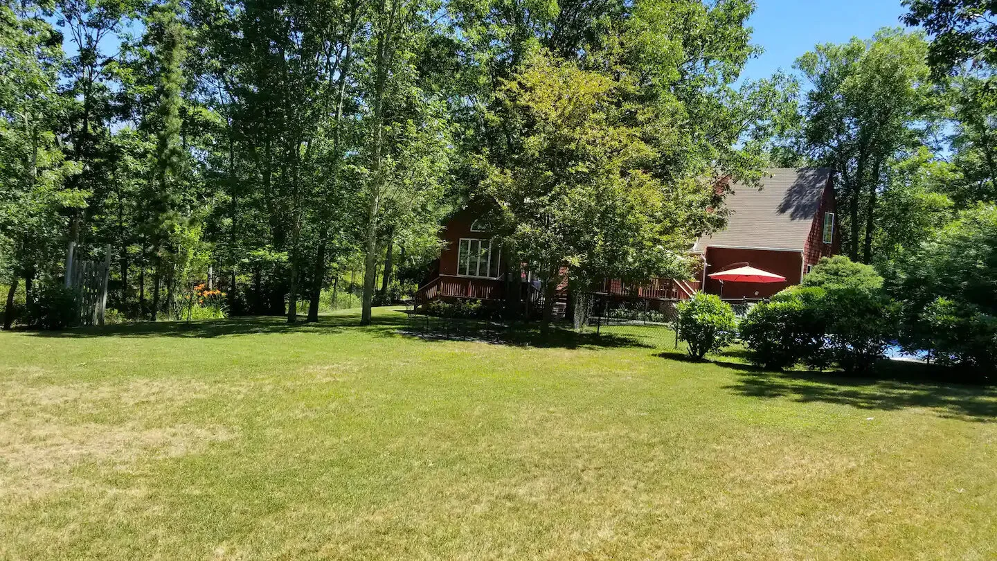 A red house stands beyond a broad grassy lawn, surrounded by trees.