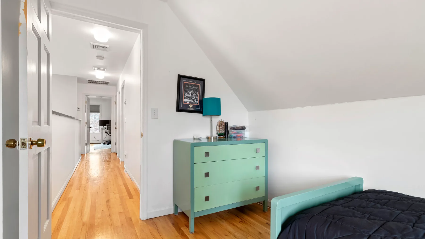 A bedroom with a bed beside a turquoise dresser and a hallway beyond.