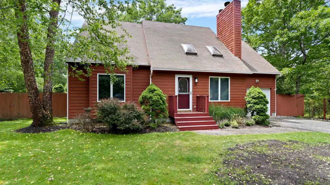 A red house stands behind a green lawn, with trees and shrubs around it.