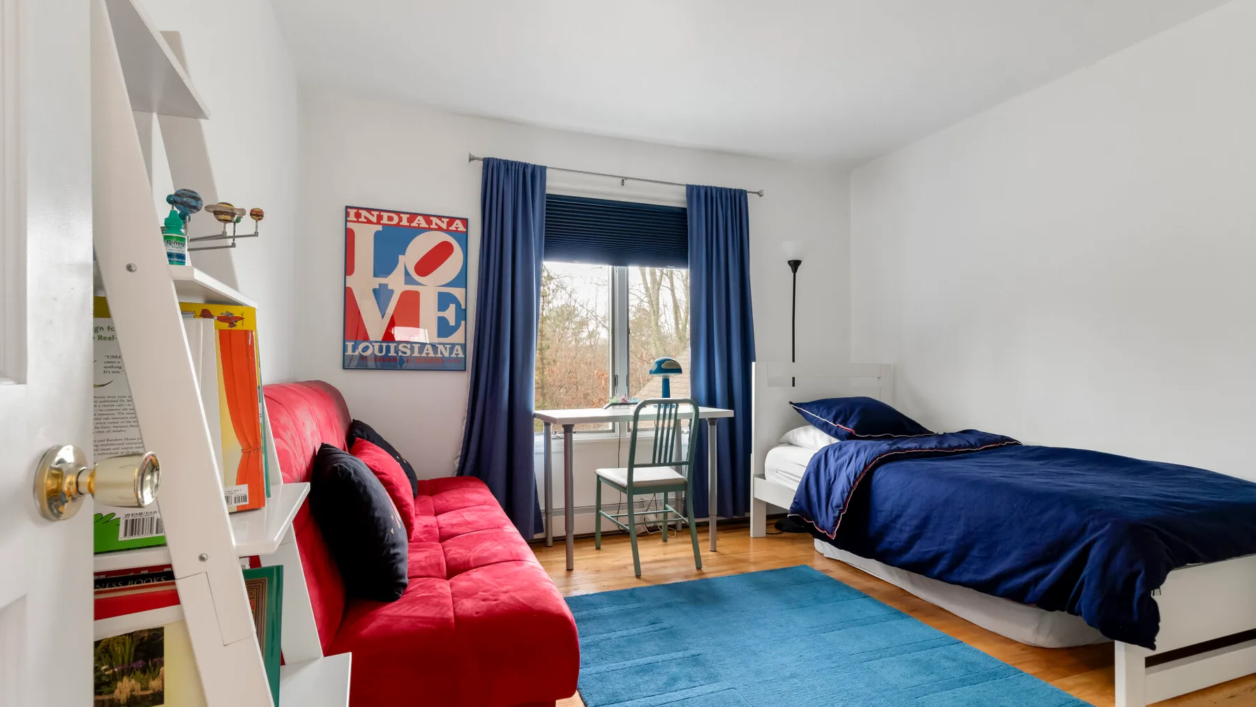 A bedroom with a red seating area, blue bedding, and a window overlooking trees.