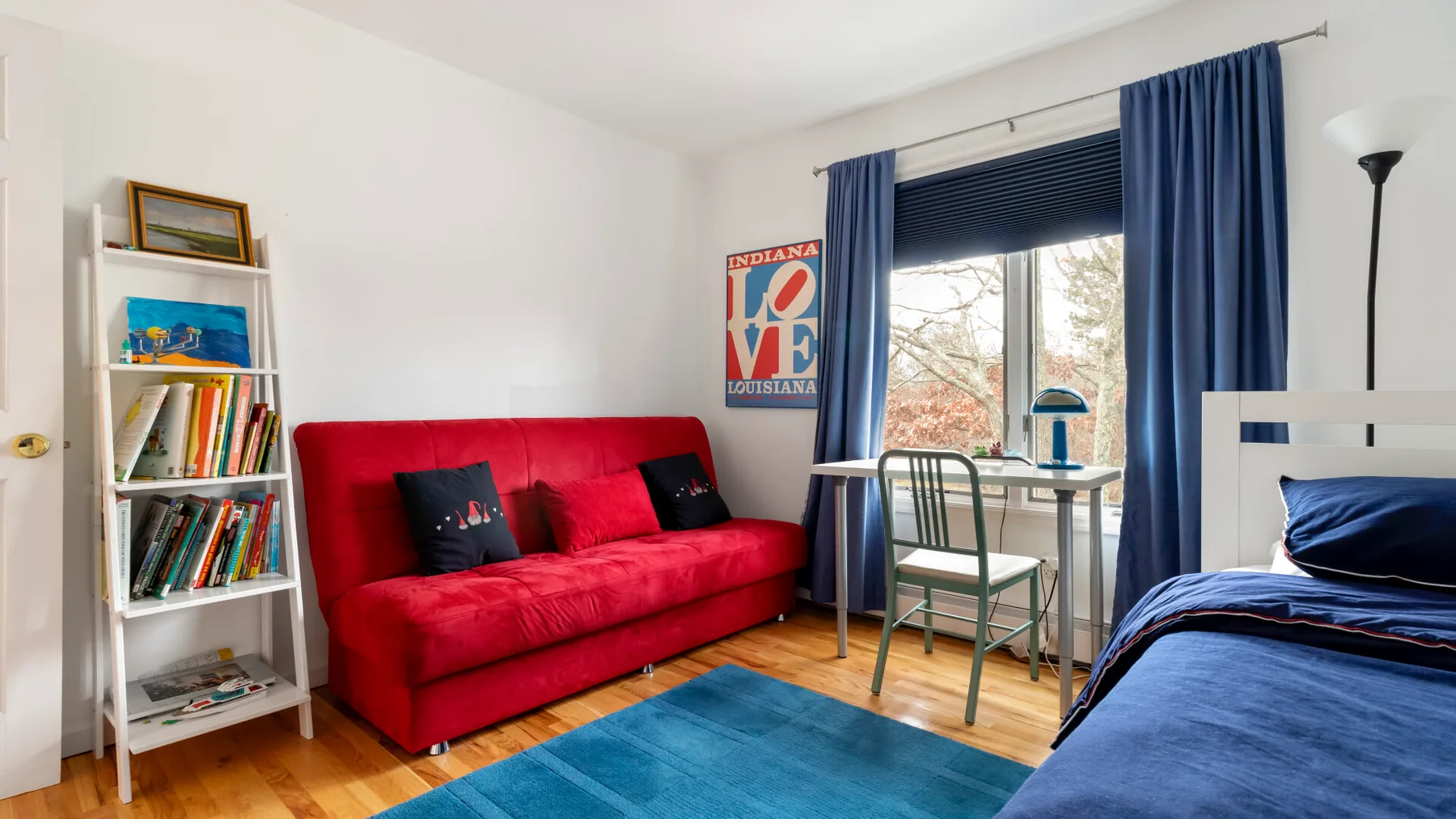 A bedroom with a red sofa, blue bedspread, and a desk beside the window.