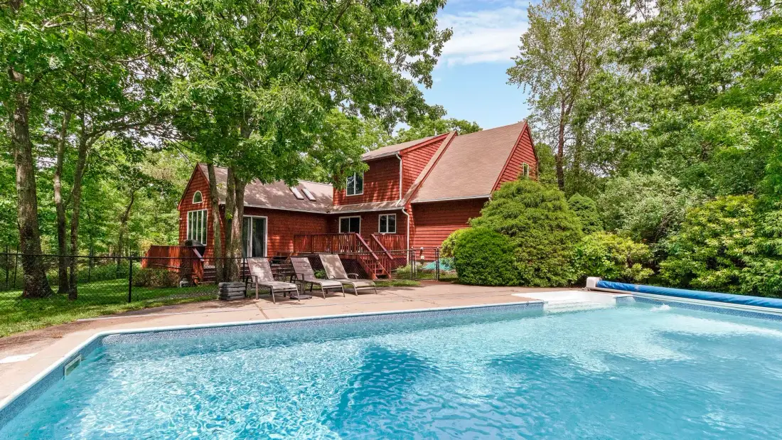 A swimming pool sits in front of a red house surrounded by trees.