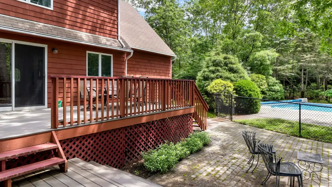 A wooden deck beside a house overlooks a fenced swimming pool and garden.