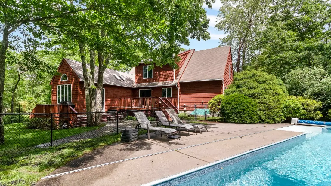 A swimming pool beside a red house, with lounge chairs on the deck.