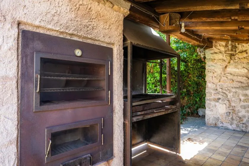 An outdoor cooking area with a built-in oven and open grill beneath a wooden roof.