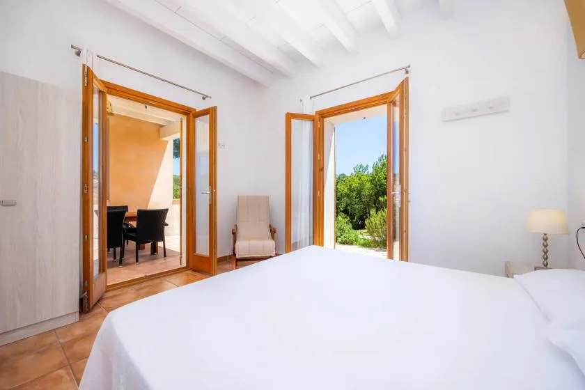 A bright bedroom has a large bed and open wooden doors leading to a garden.