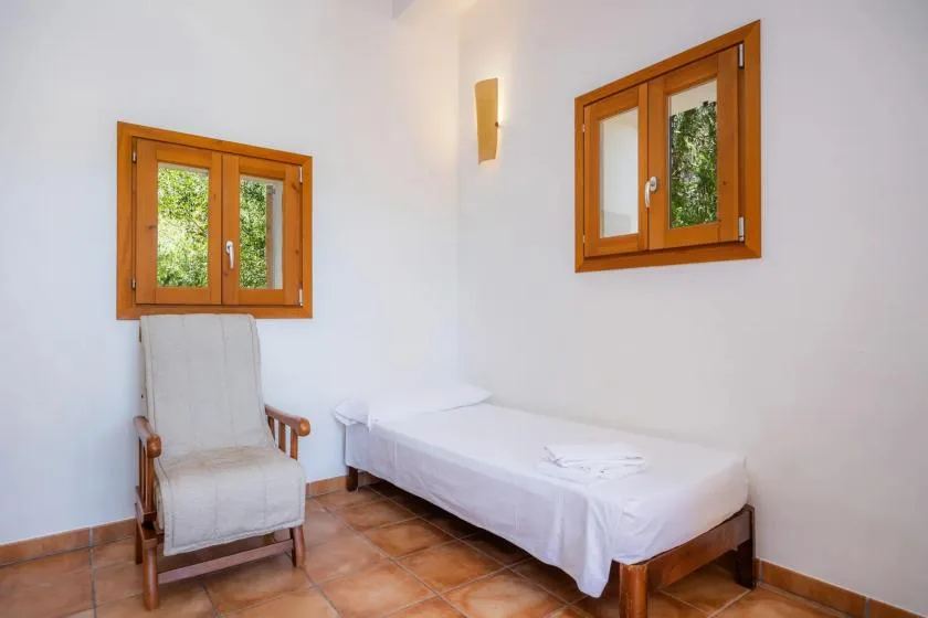A simple bedroom with a single bed and a cushioned chair beneath two windows.