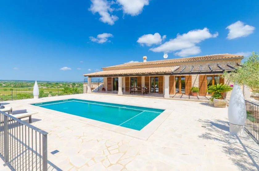 A sunlit swimming pool and paved patio sit beside a low villa.