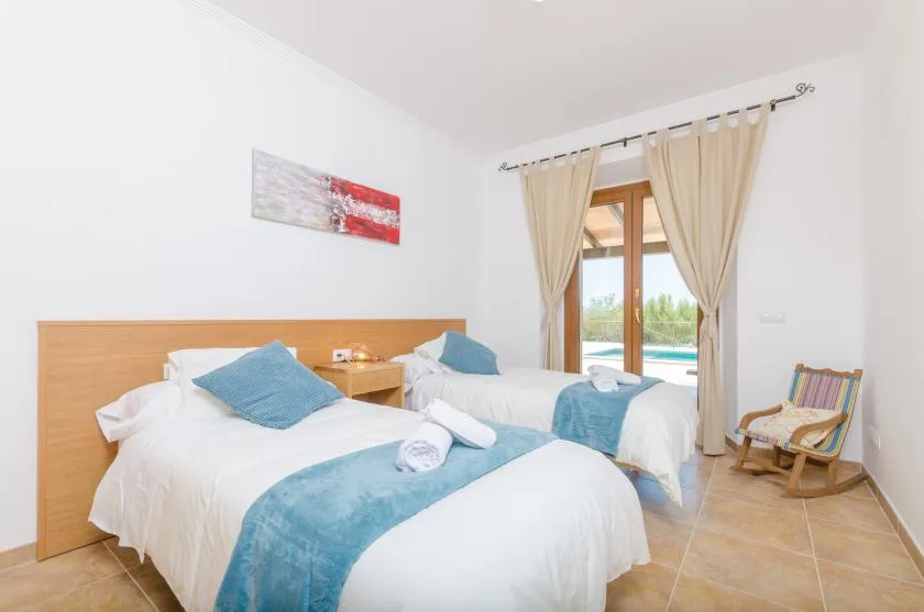 A bright bedroom with two beds, blue bedding, and a window with curtains.
