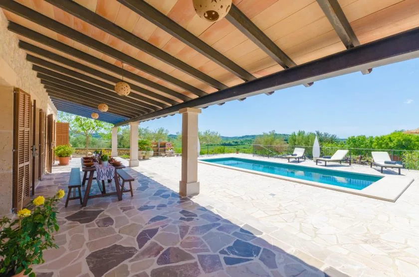A covered patio with outdoor seating overlooks a swimming pool and green landscape.