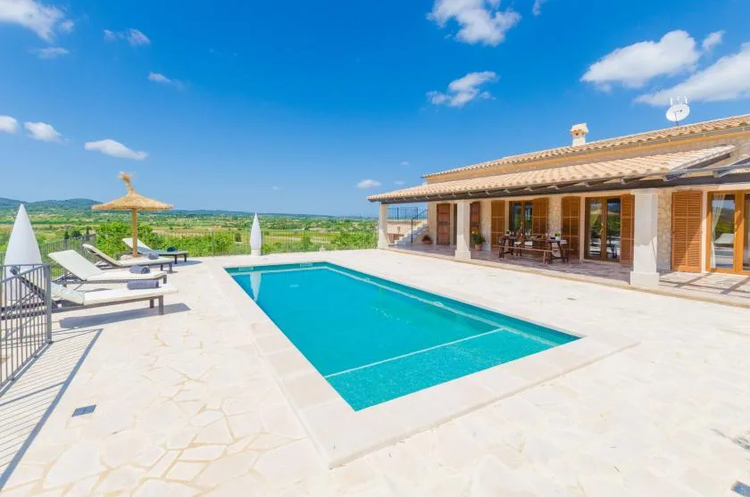 A sunny outdoor pool patio beside a house, with lounge seating and open countryside views.