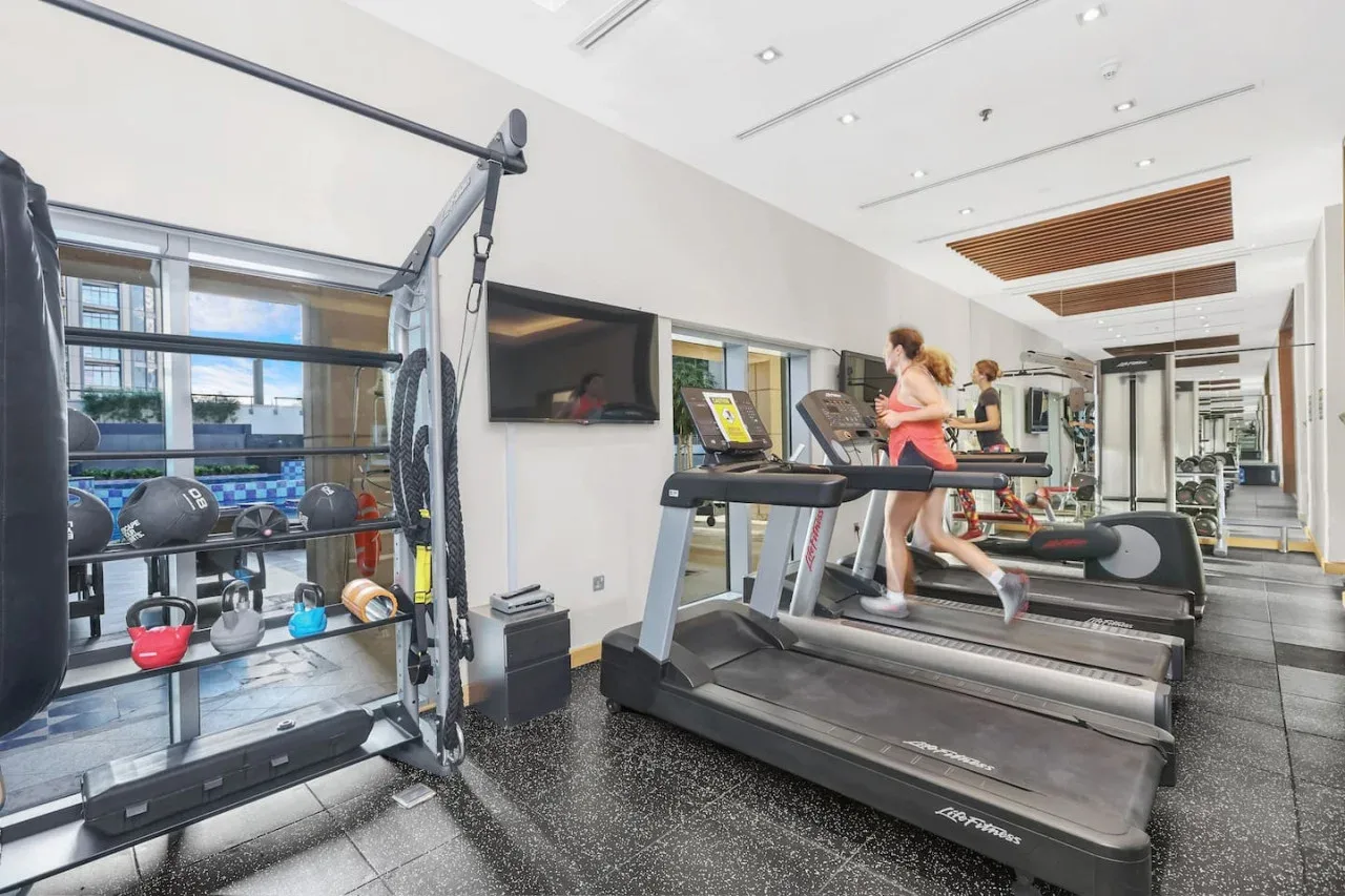 Un gimnasio con cintas de correr y equipo de ejercicio junto a grandes ventanas.