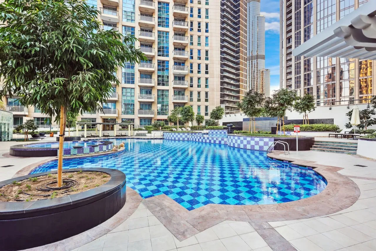 Una piscina al aire libre con azulejos azules está rodeada de jardines y edificios altos.