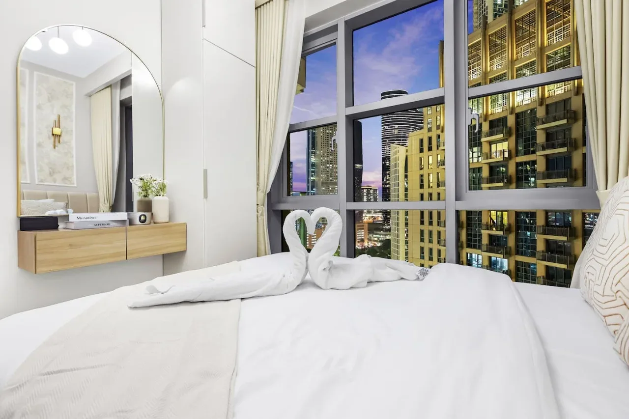 Un dormitorio luminoso con ropa de cama blanca y una gran ventana con vistas a edificios de la ciudad.