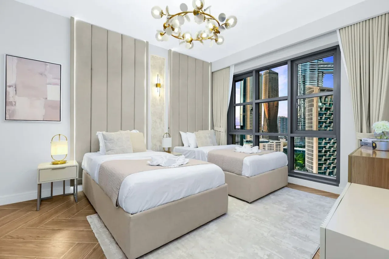 Un dormitorio con dos camas, muebles de tonos neutros y grandes ventanas con edificios a la vista.