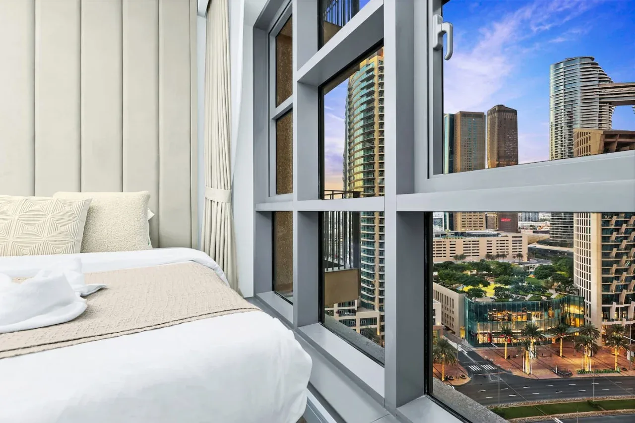Un dormitorio con una cama junto a grandes ventanas con vistas a edificios de la ciudad.