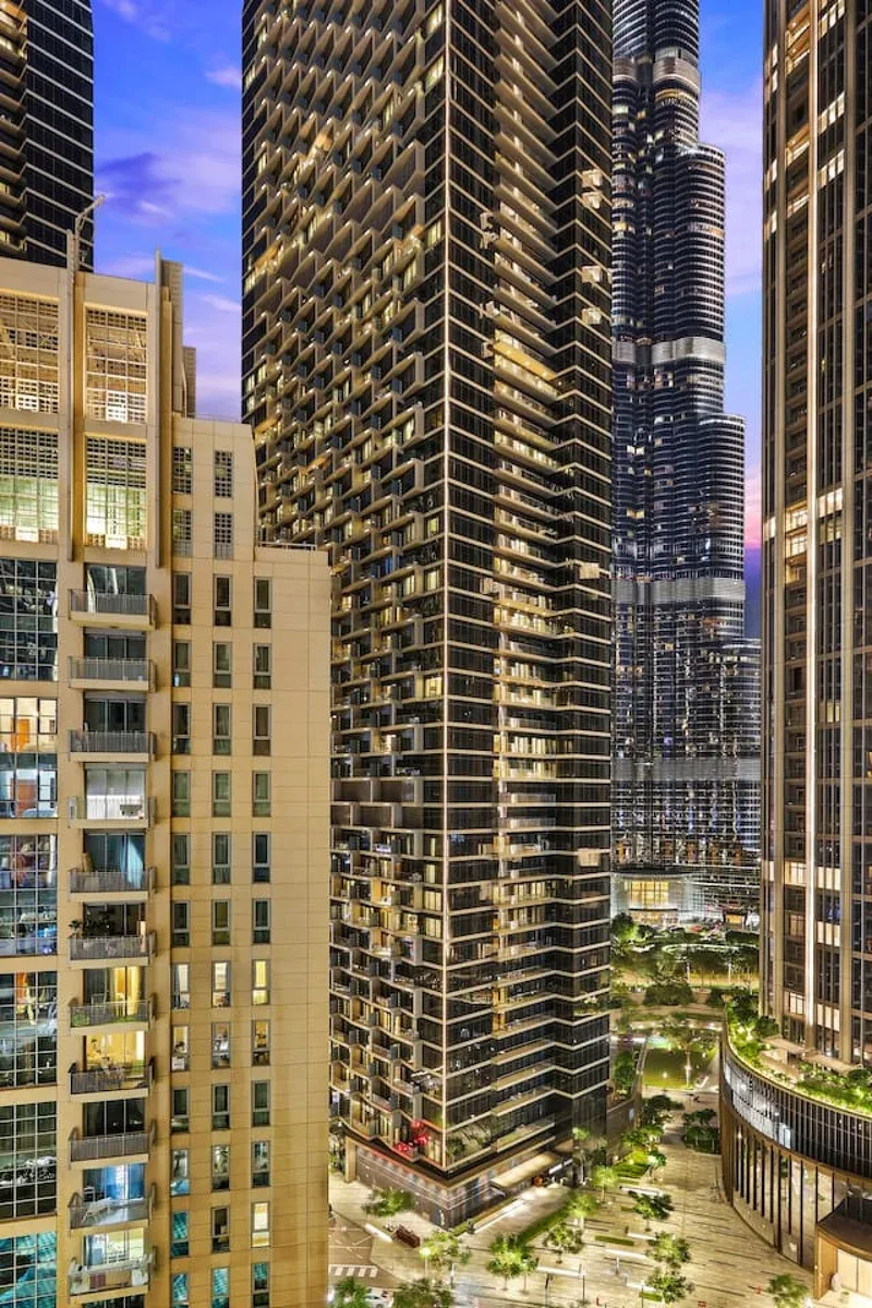 Altos edificios urbanos se alzan sobre una plaza ajardinada bajo un cielo azul al anochecer.