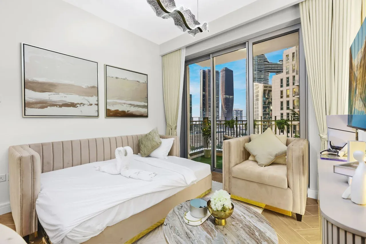 Un dormitorio luminoso con cama, asientos y grandes ventanas con vistas a edificios cercanos.