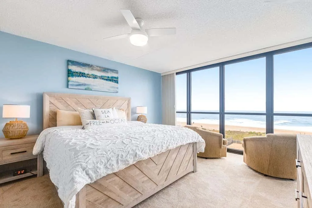 A bedroom with a bed beside large windows overlooking the beach.