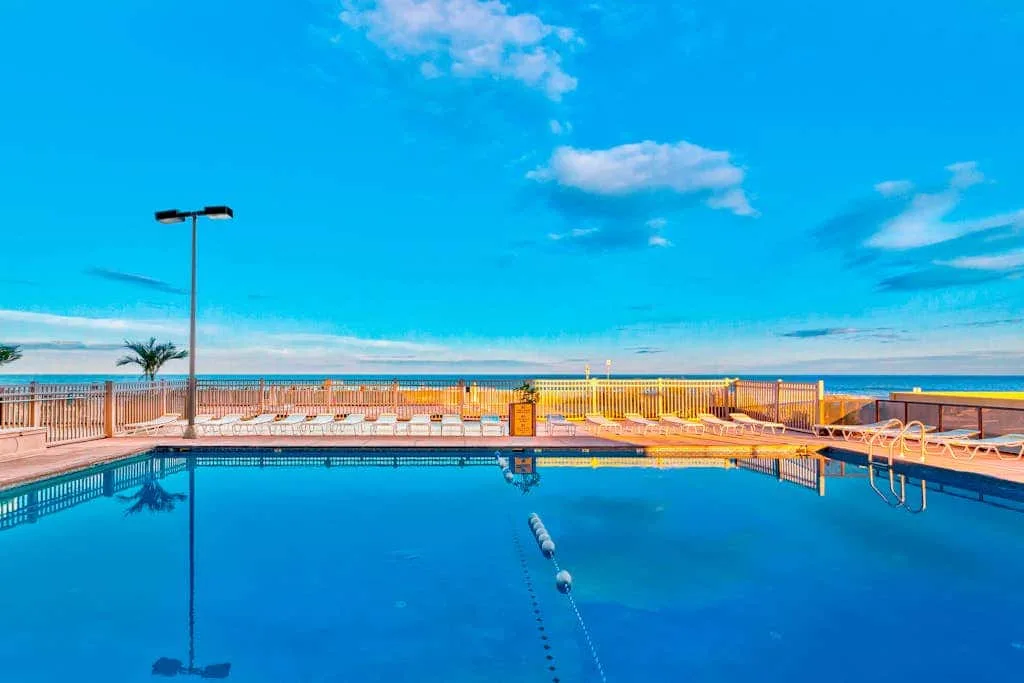 An outdoor swimming pool overlooks the ocean beneath a bright blue sky.