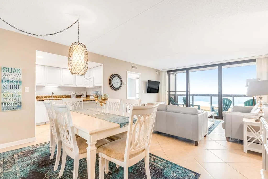 An open dining and living area has a table beside large windows overlooking the water.