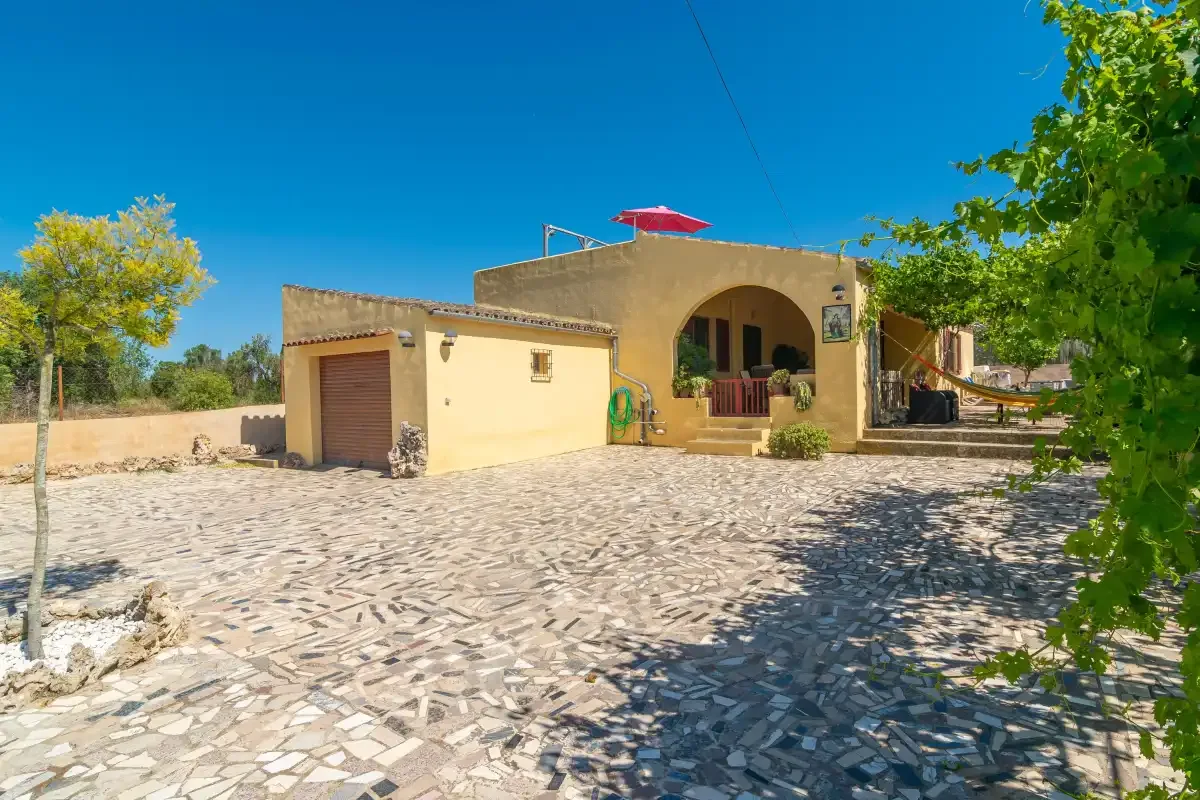 Una casa amarilla se alza junto a un amplio patio pavimentado bajo un cielo azul despejado.