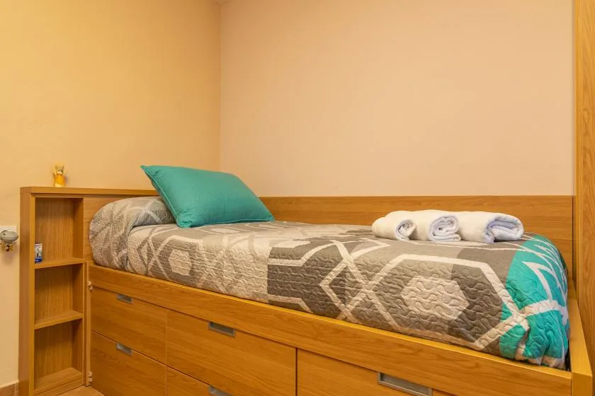 Una cama individual con colcha estampada y almohada turquesa está junto a una pared lisa.