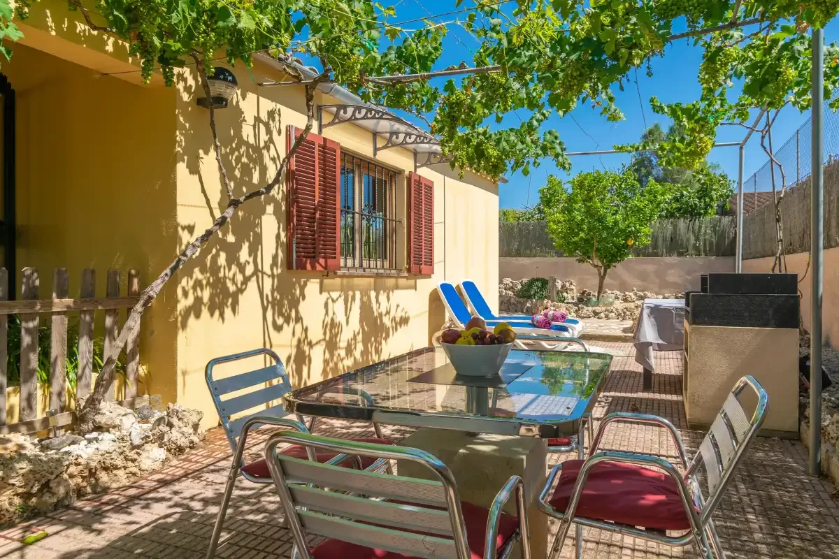 Una zona de comedor exterior a la sombra junto a una casa amarilla, con mesa y asientos.