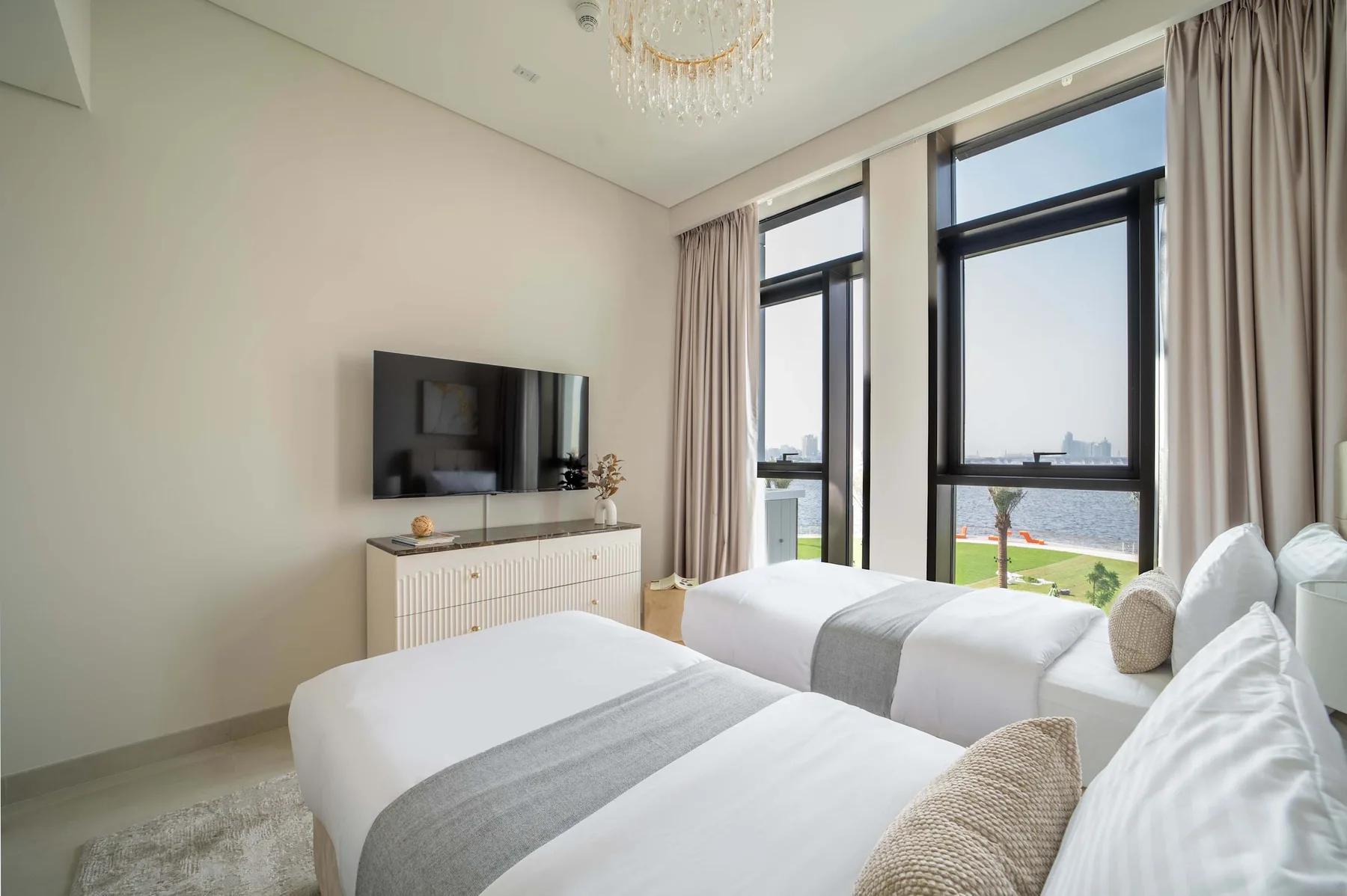 Un dormitorio luminoso con dos camas, un televisor en la pared y grandes ventanas.