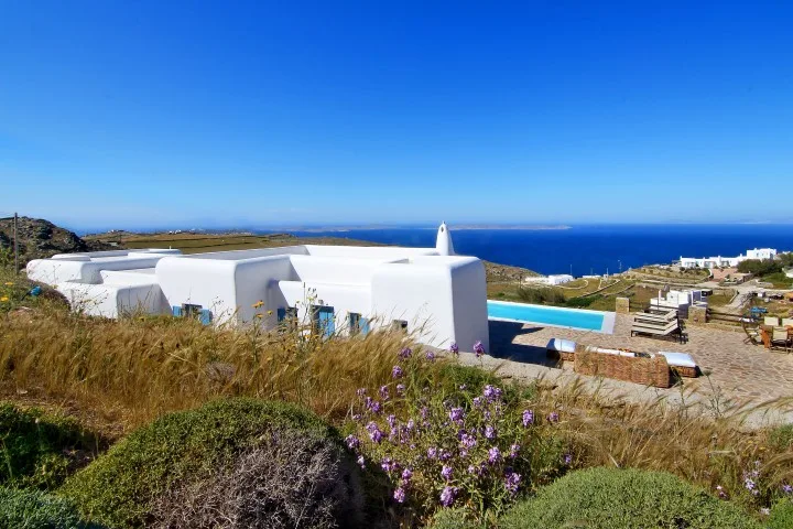 White buildings and a turquoise pool overlook the sea on a sunny hillside.