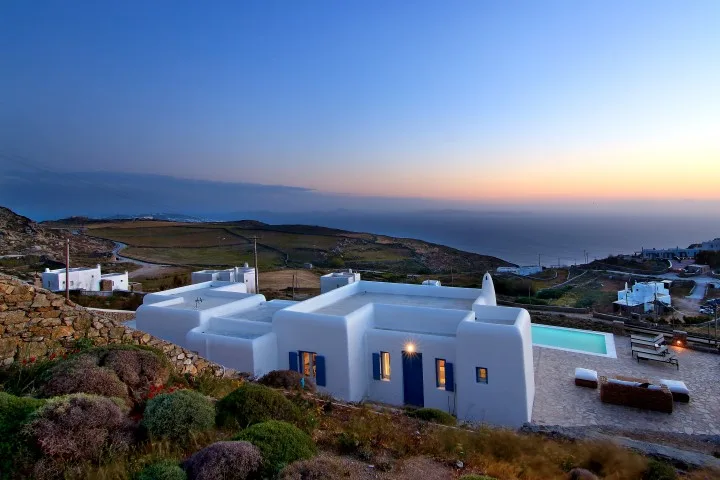 White buildings with a pool sit among rocky hills overlooking the sea at dusk.
