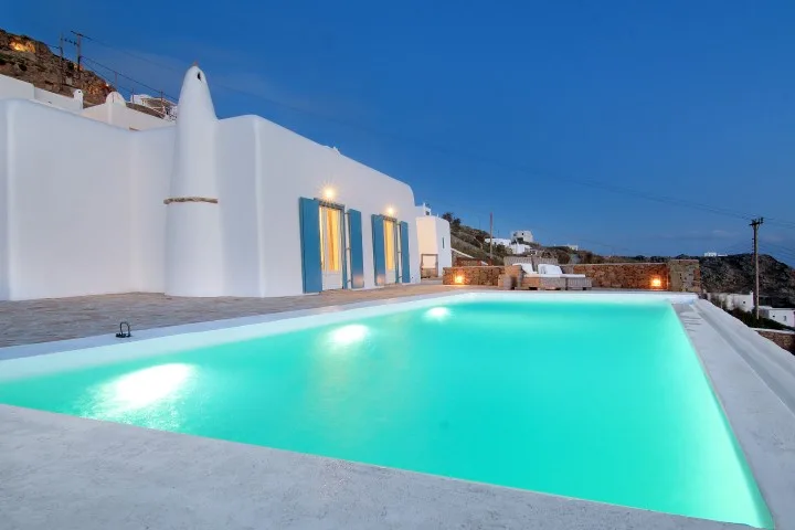 An outdoor swimming pool beside white walls and glass doors at dusk.