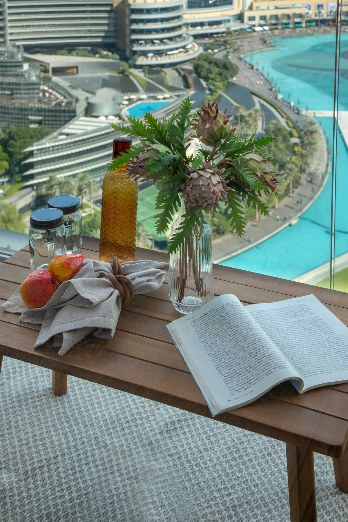 Una mesa junto a una ventana grande tiene un libro abierto, fruta y un arreglo floral.