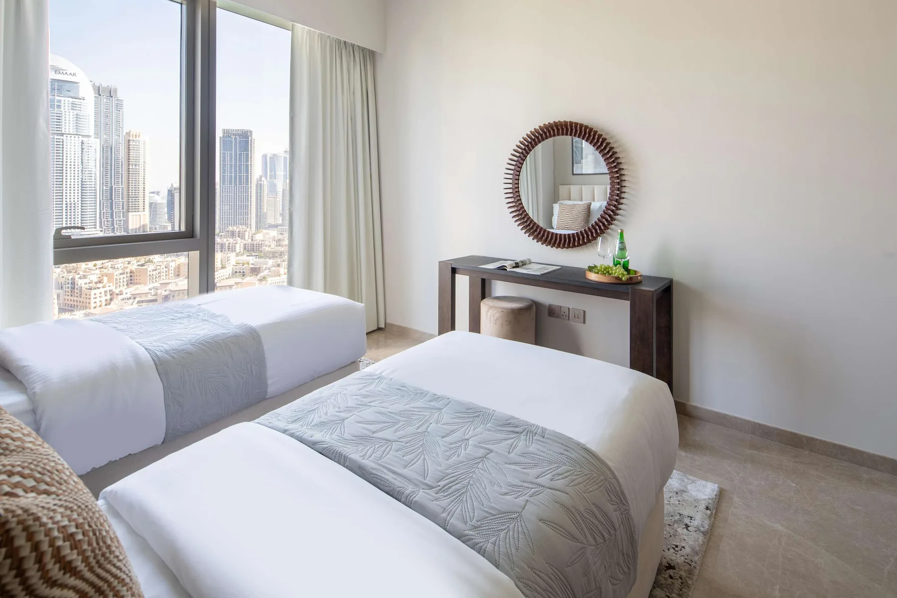 Un dormitorio luminoso con dos camas junto a una ventana amplia con vistas a edificios.