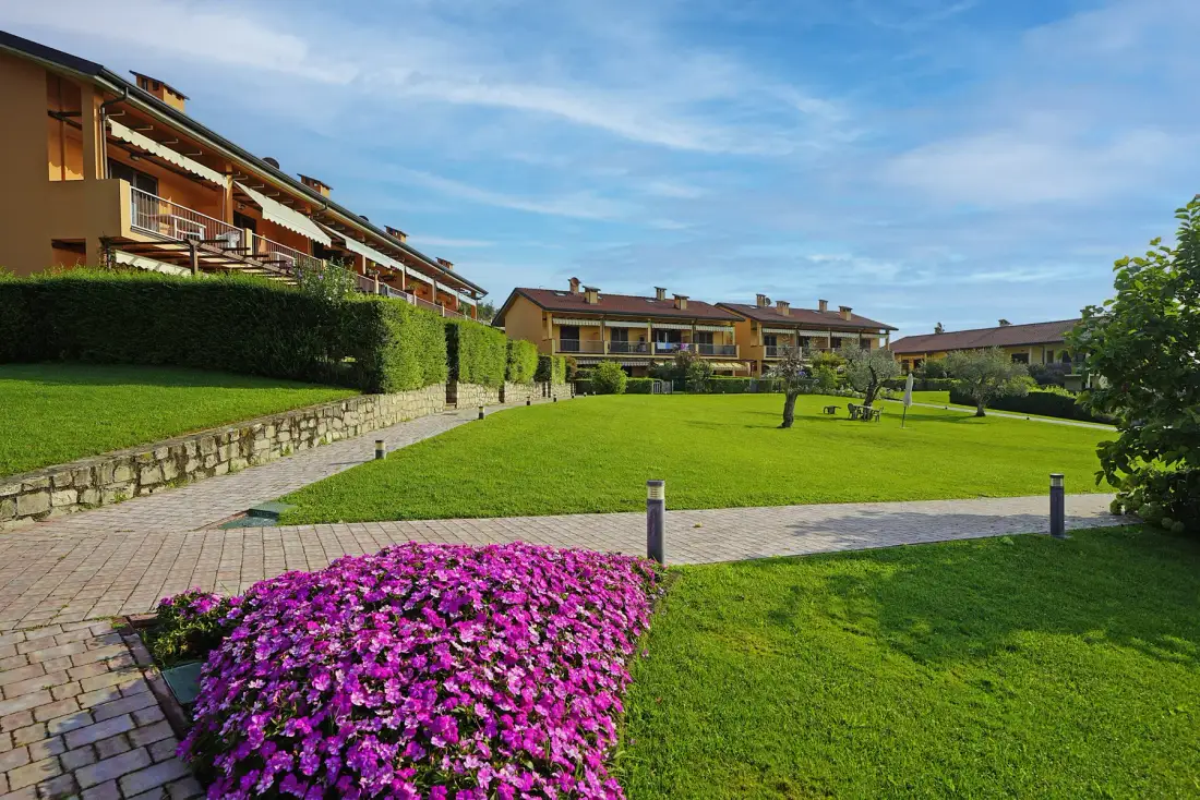 Residential buildings overlook a green lawn with paved paths and flowering plants.