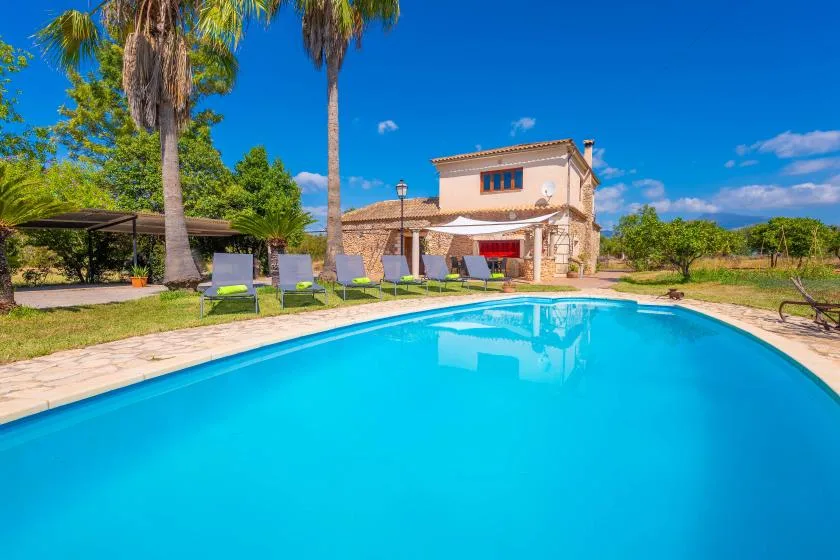 Una piscina junto a una casa de piedra y asientos al aire libre bajo palmeras.